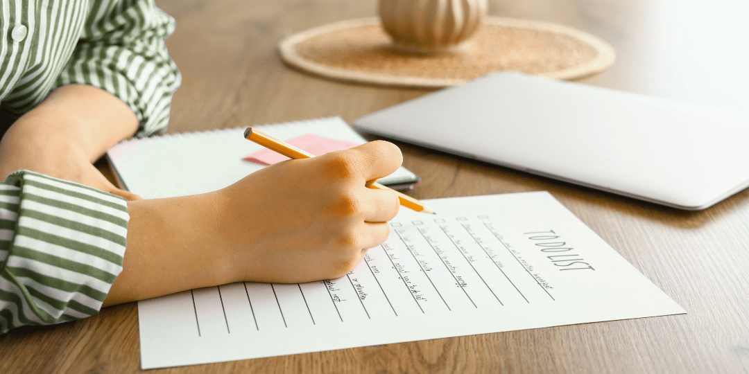 A person sitting at a wooden table in a brightly lite room wearing a green and white striped shirt. They have a pencil in their hand and appear to be writing a to-do list showing an example of the additional services that Ebb & Spruce offers. 