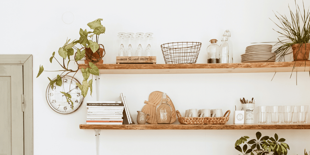 An organized shelf in a brightly lit room with neatly arranged green plants, glasses, and cook books. Everything has it's place a beautiful example of how Ebb & Spruce can help you get organized.  