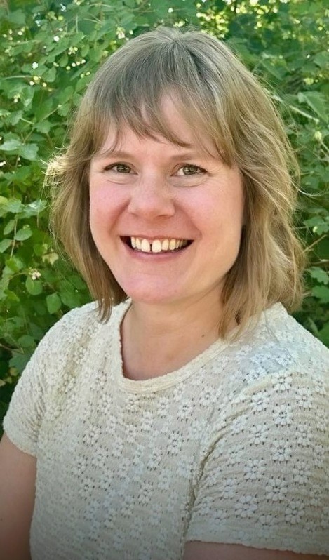 Sarah Tudway-Cains, the owner of Ebb & Spruce is standing outside on a sunny day in front of green foliage. She loves working with clients from all different backgrounds, and helping them create the spaces they can thrive in.